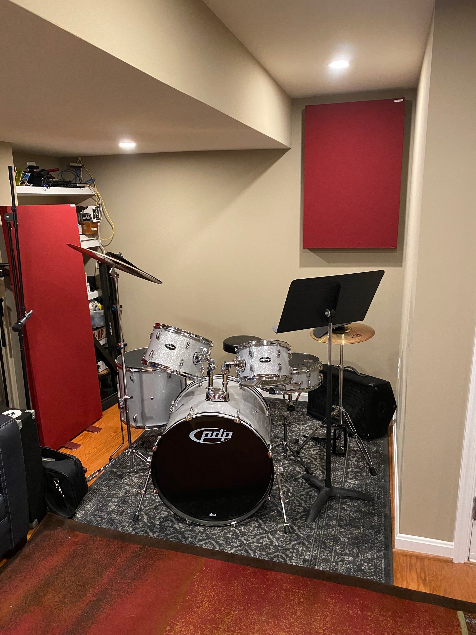 A silver drum kit on a rug in an alcove with a red acoustic panel on the wall and a music stand beside it