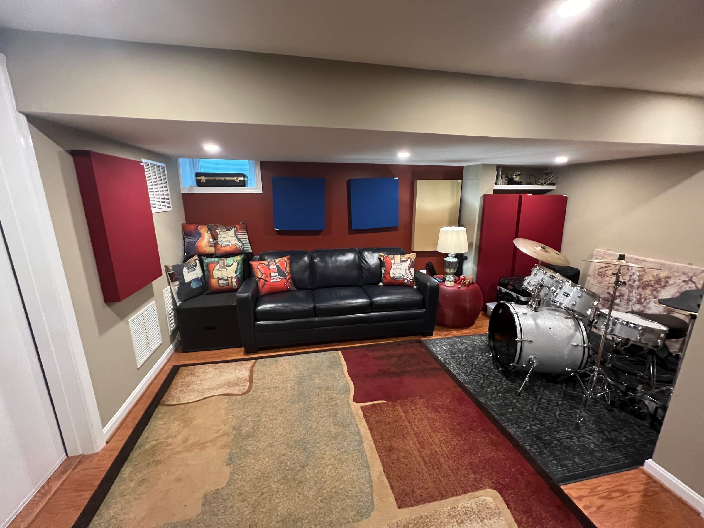 The studio lounge: a black leather sofa with guitar-print pillows, red and blue acoustic panels on the walls, and a silver drum kit on a rug to the right